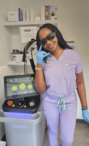 Dr. Lian Peter dans sa tenue violette, souriant et posant à côté du système laser Cynosure Elite IQ à l'intérieur de la salle de procédure de la clinique d'esthétique médicale Dr. Lian Beauty à Limoges, en Ontario.