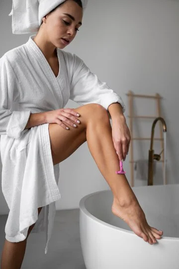 Woman in a white bathrobe shaving her leg raised on a bathtub